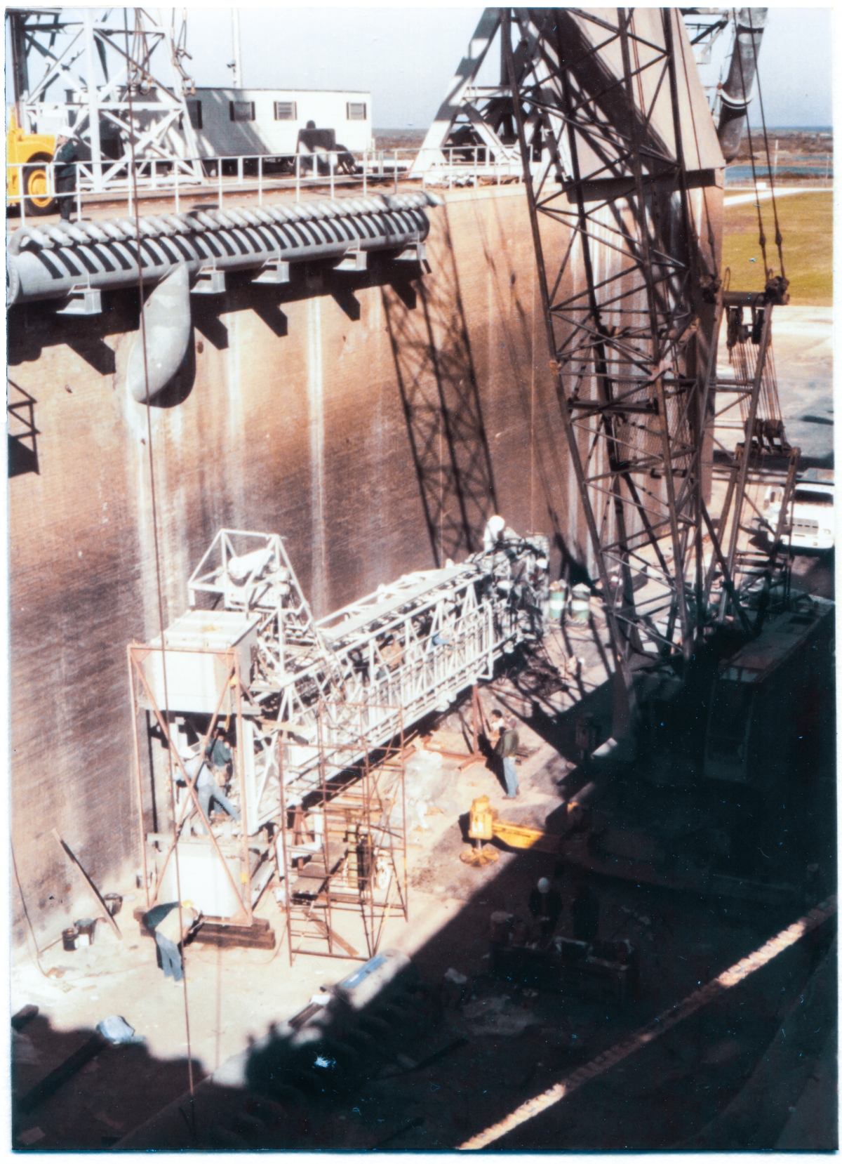 Image 129. At Space Shuttle Launch Complex 39-B, Kennedy Space Center, Florida, preparatory work on the Orbiter Access Arm (OAA) is in progress, prior to it being hung on the tower. The OAA is sitting on metal support stanchions, down in the bottom of the Flame Trench, north of the Flame Deflector. This is the same location that work on the GOX Arm was done, prior to it getting lifted. From here, it's pretty straightforward crane work to pick it up and place it on the Pad Deck, where the final preparations will be completed, immediately prior to lifting the Arm, and hanging it on Side 1 of the FSS at elevation 200'-0”. At the time this photograph was taken, no sensible structural work was being done on the Arm, and instead, it was all Electrical and Mechanical trades work. The Environmental Chamber (also known as the ”White Room”) has yet to be attached to the far end of the arm, opposite along its length, away from the Hinges, which attach directly to the Strongback on the FSS, and which can be seen in the lower left portion of this image. Photo by James MacLaren.